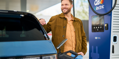 Un homme sourit à côté de sa voiture et de la borne de recharge Q8 electric pendant la recharge.